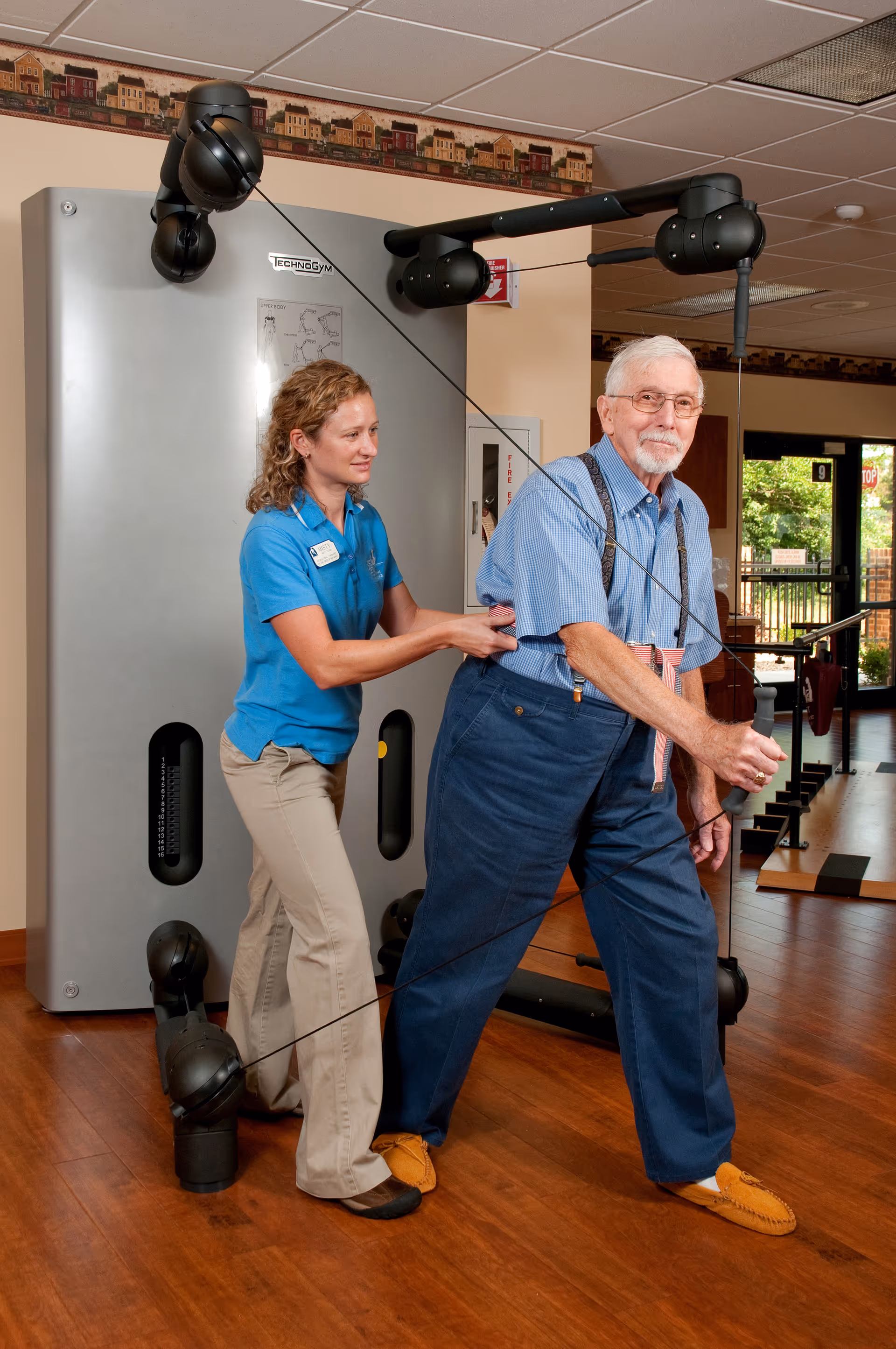 An elderly man is exercising with resistance cables on a fitness machine while a female staff member assists him in a well-lit room with wooden floors and large windows.