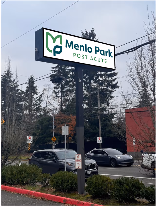 Outdoor view of a tall signpost with a rectangular sign displaying the logo and name 'Menlo Park Post Acute' in green and black text. The sign is situated near a road with cars passing by, surrounded by trees and bushes under an overcast sky.