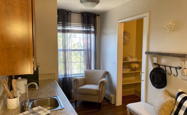 Small kitchenette with a countertop and sink, an upholstered armchair by a curtained window, and an open pantry doorway.