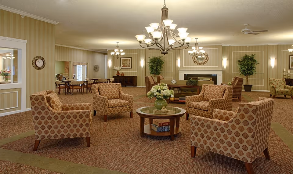 A spacious and well-lit living room area in a senior living facility with patterned armchairs arranged around a round glass-top coffee table holding a green vase with flowers. The room features beige striped wallpaper, a carpeted floor, multiple chandeliers, a fireplace with a decorative mirror above it, and potted plants. In the background, there are dining tables and chairs.