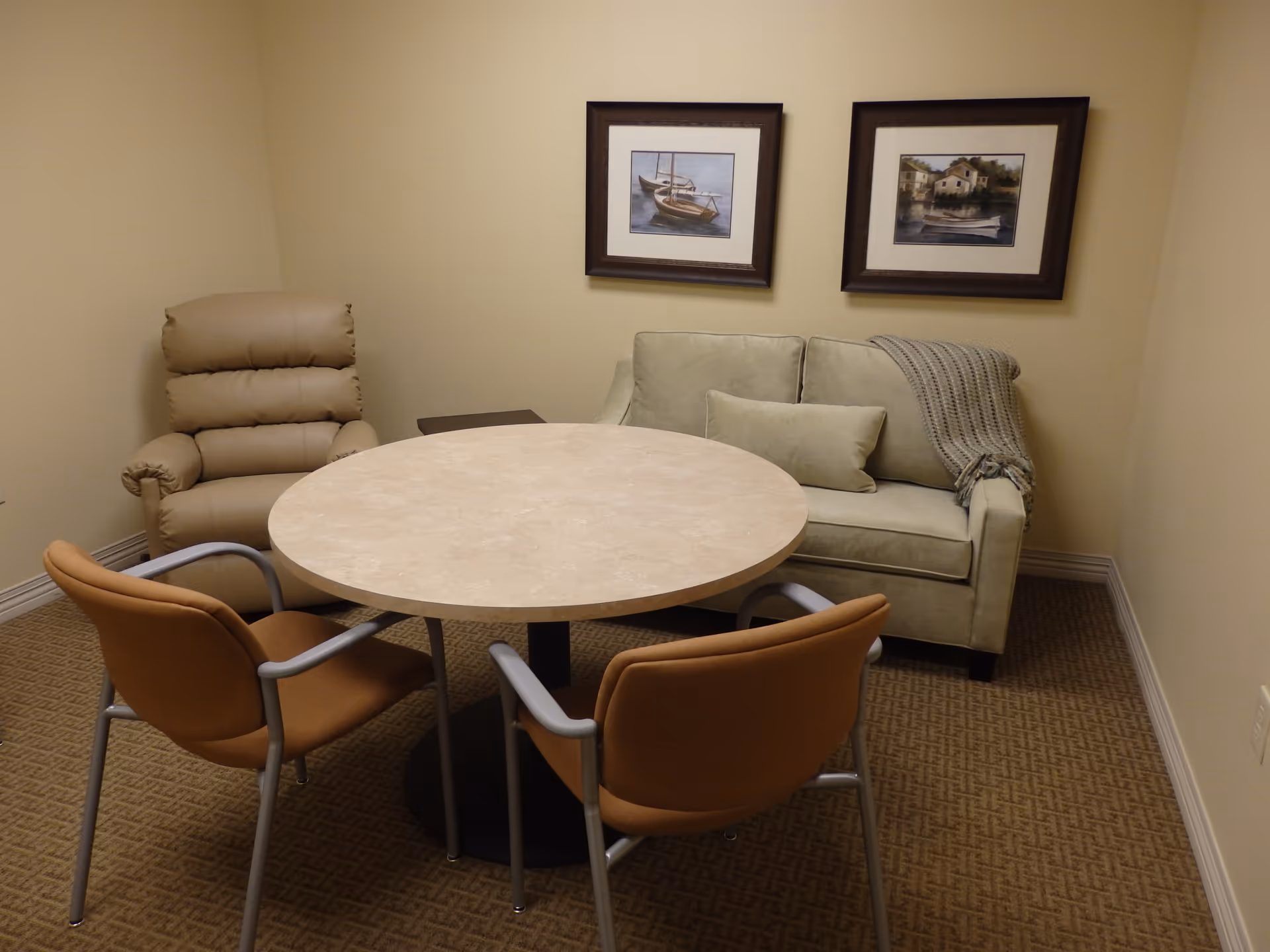 Small room with a round table surrounded by two brown chairs, a beige recliner, and a light green loveseat with a throw blanket and pillow. Two framed pictures of boats and houses hang on the beige wall above the loveseat.