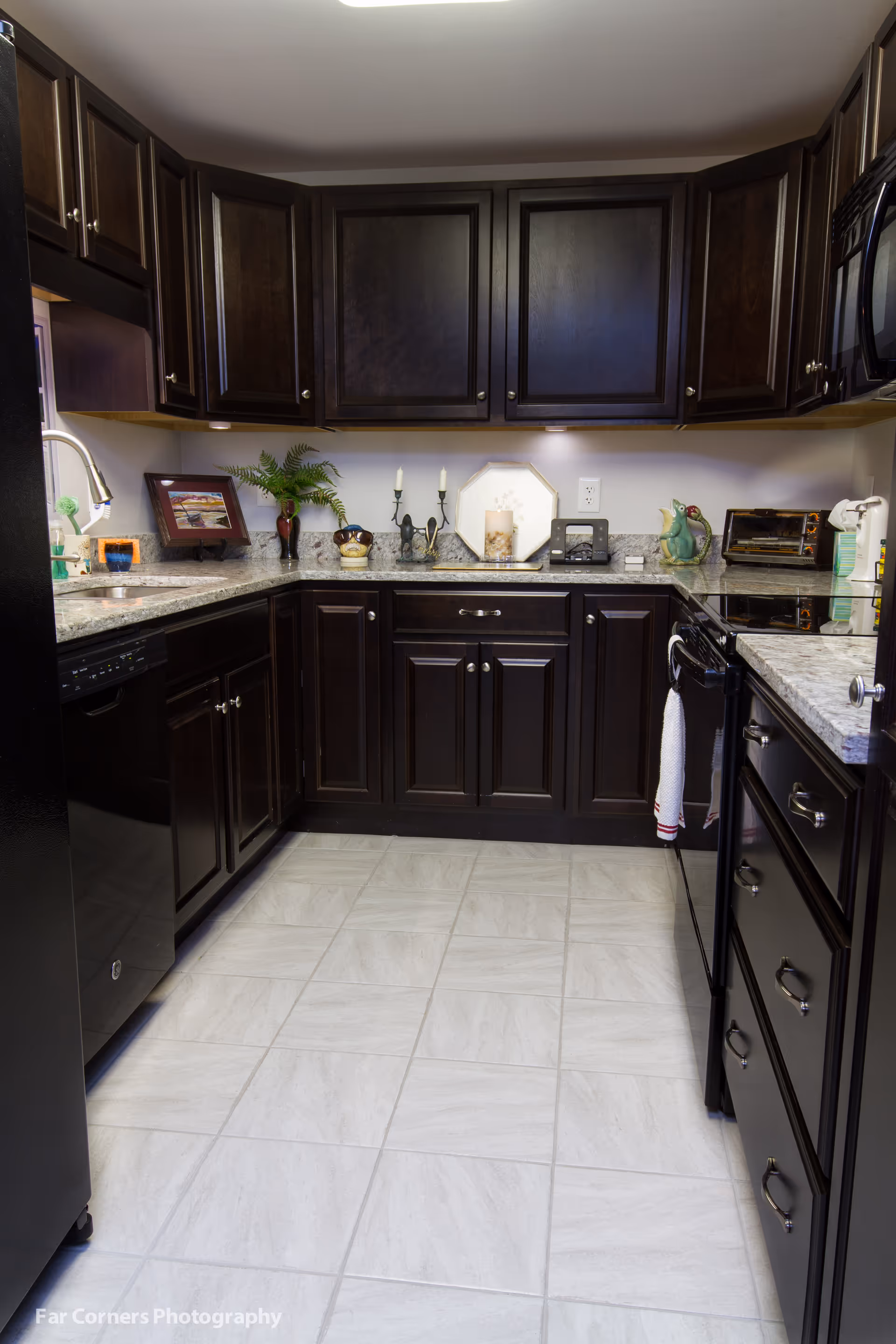 A modern kitchen with dark wooden cabinets, light-colored granite countertops, and light gray tiled flooring. The kitchen features a black dishwasher, black stove with a white towel hanging on the handle, a microwave, a toaster oven, and various decorative items including a plant, candles, and framed pictures.