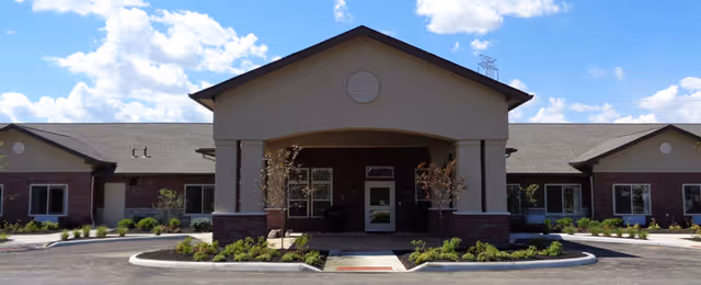 Front exterior view of a single-story assisted living and memory care facility building with a covered entrance, landscaped greenery, and a clear blue sky with some clouds.