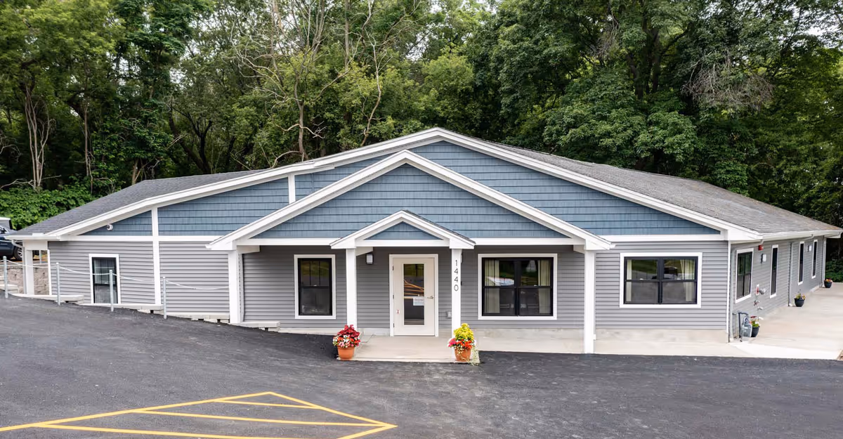 Front exterior view of a single-story assisted living facility building with gray siding and white trim, a small covered entrance with two potted plants, and surrounded by trees in the background.