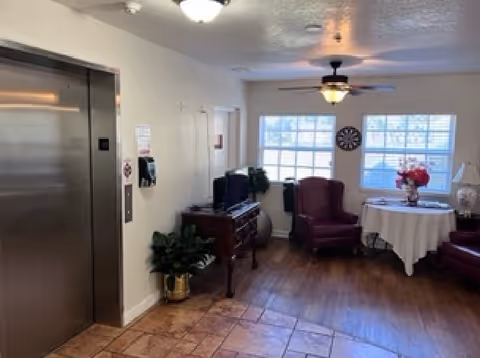 Interior view of a senior living facility common area with an elevator on the left, a small wooden cabinet with a TV, two armchairs, a round table with a white tablecloth and a flower vase, and two windows letting in natural light.