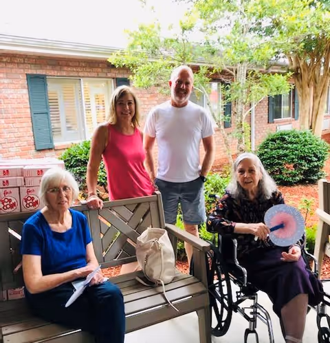 Two elderly women and two middle-aged adults are outside a brick building with green shutters. One elderly woman is sitting on a bench wearing a blue shirt, and the other elderly woman is in a wheelchair holding a pink and blue fan. A woman in a pink sleeveless top and a man in a white t-shirt and shorts stand behind them. There are bushes and trees in the background.