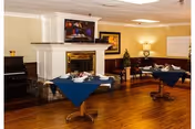 Interior view of a common area in a senior living facility with wooden flooring, two tables covered with blue tablecloths and set with dining ware, a fireplace with a television mounted above it, a piano on the left, and framed artwork on the walls.