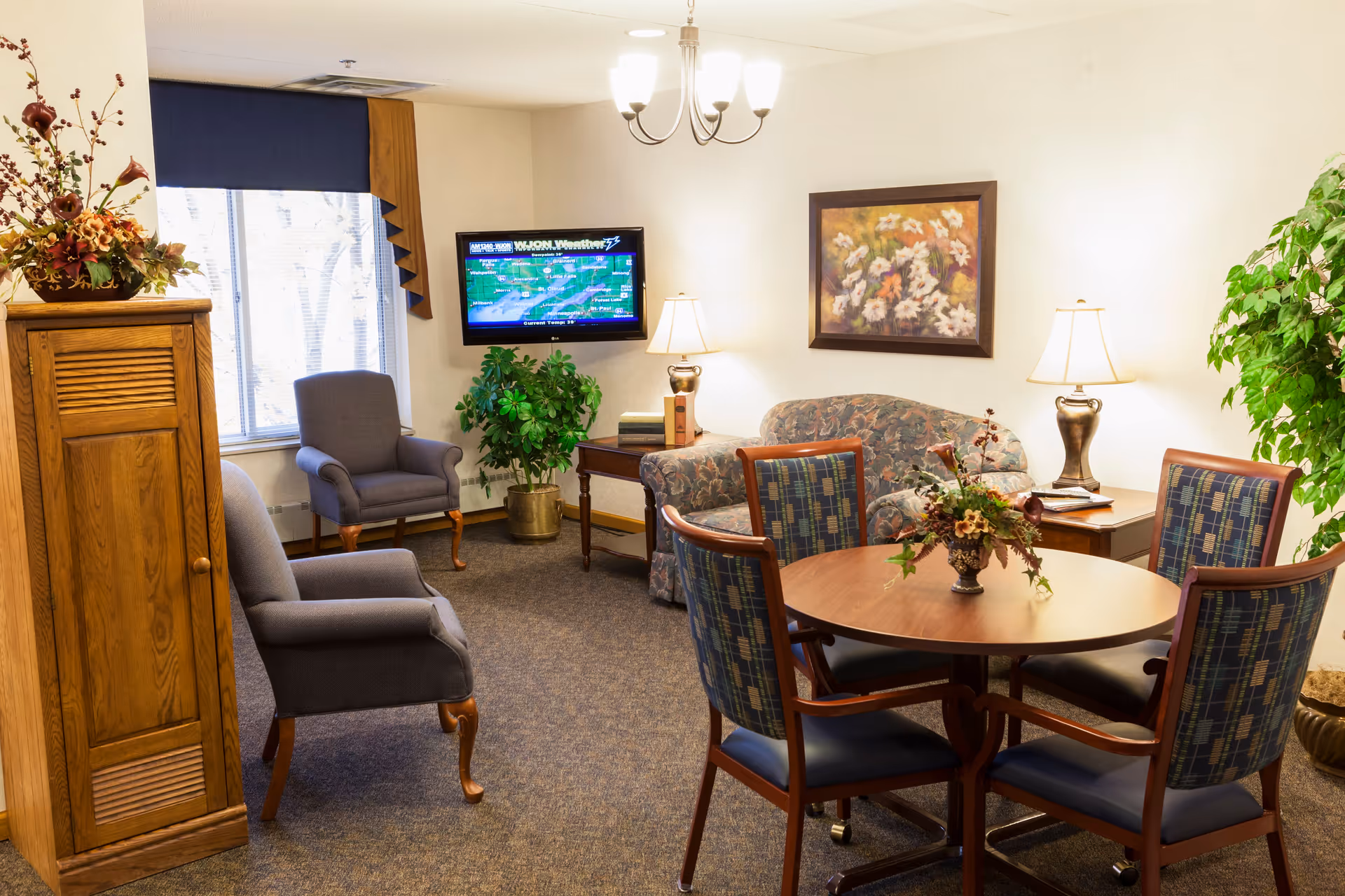 A cozy living room area with a round wooden table surrounded by four cushioned chairs, a floral arrangement centerpiece on the table, two upholstered armchairs, a floral-patterned sofa, two table lamps, a wall-mounted TV displaying a weather map, a wooden cabinet, and potted plants near the window and corner.