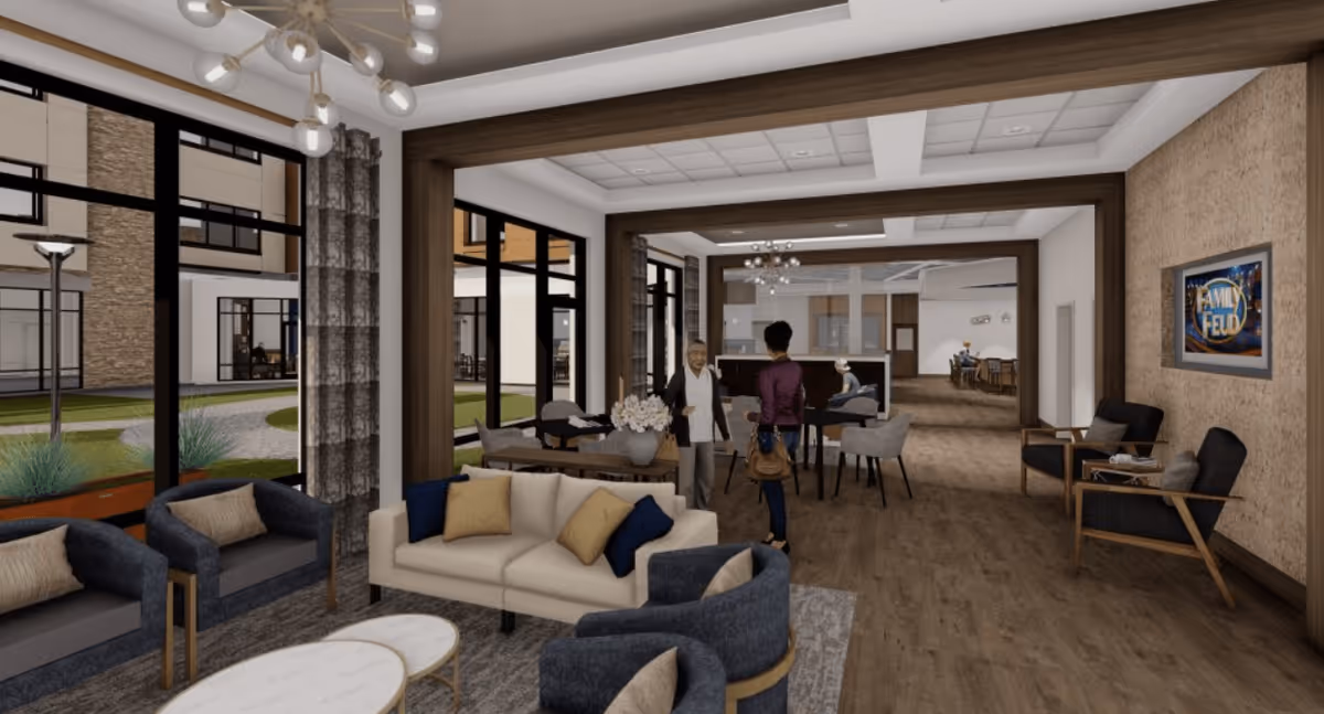 A spacious and modern senior living facility common area with comfortable seating including sofas and armchairs arranged around a coffee table. Large windows provide a view of an outdoor courtyard. Two people are standing and conversing near a dining table in the background. The room features wood flooring, contemporary lighting fixtures, and a framed 'Family Feud' picture on the wall.