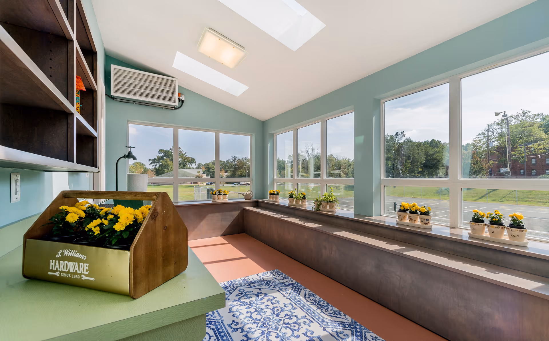 A bright sunroom with large windows and skylights letting in natural light. The room has a green countertop with a wooden planter box labeled 'S. Williams Hardware' containing yellow flowers. Along the windowsills are multiple small pots with yellow flowers and green plants. Outside the windows, a grassy area and trees are visible.