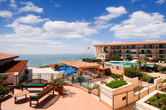 Outdoor patio area at White Sands La Jolla with wooden benches, potted plants, a pergola with seating underneath, blue umbrellas, a swimming pool, and a multi-story building in the background overlooking the ocean under a partly cloudy sky.