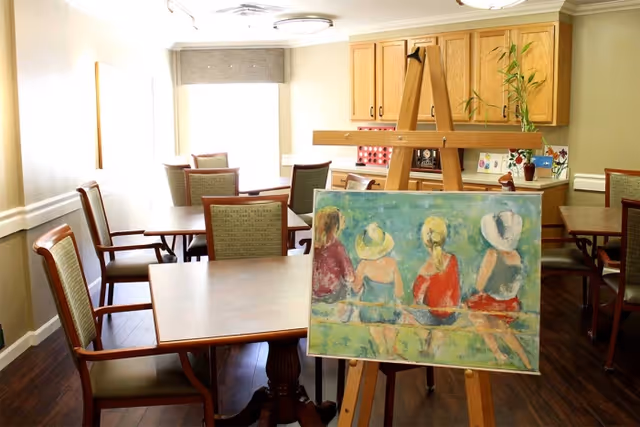 A well-lit room with several wooden tables and chairs arranged neatly. In the foreground, there is an easel displaying a colorful painting of four women sitting with their backs to the viewer, wearing hats and summer clothing. The room has wooden flooring, light-colored walls, and a window with a gray valance letting in natural light. There are wooden cabinets and some decorative items on a counter in the background.
