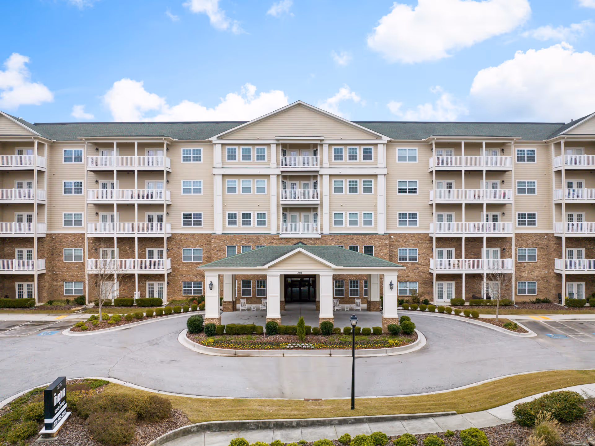 Front exterior view of a large senior living facility building with multiple floors, balconies, and a covered entrance driveway. The building has beige siding with brick accents and a green roof. There is a landscaped area with shrubs and flowers in front of the entrance, and a sign near the driveway.
