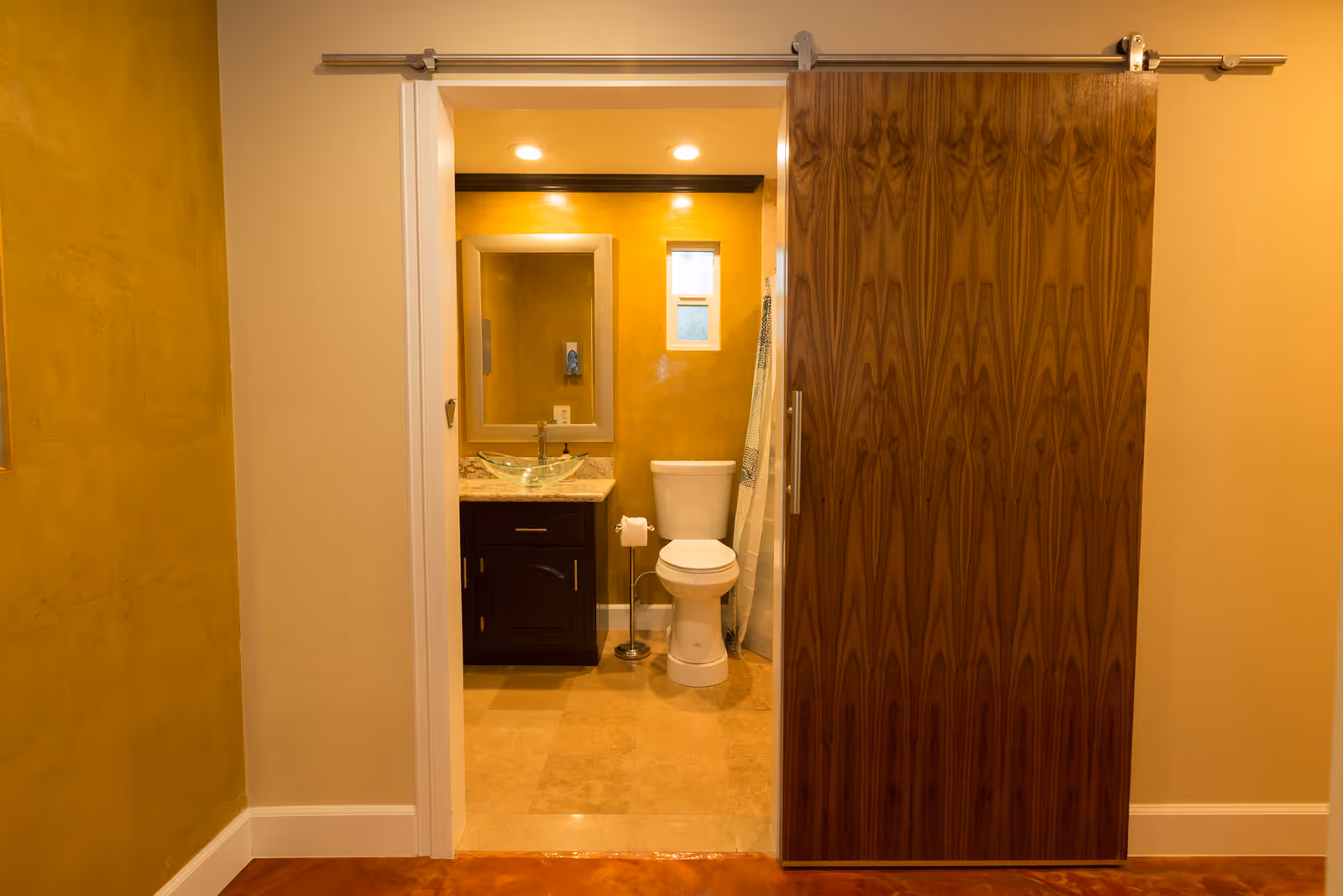 View of a bathroom with a sliding wooden door partially open. Inside the bathroom, there is a toilet, a dark wooden vanity with a glass vessel sink, a large mirror above the sink, and a small window. The walls are painted a warm yellow color and the floor is tiled.