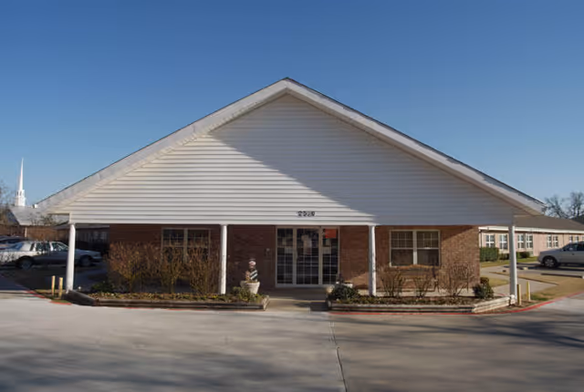 Front entrance of a single-story brick building with white siding gable, covered porch supported by columns, and a driveway.
