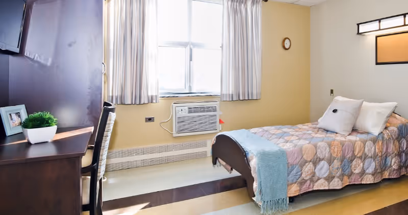 A bright and clean bedroom in a senior living facility featuring a single bed with a colorful quilt and pillows, a wooden desk with a chair, a small potted plant, a framed photo, a wall-mounted TV, a window with curtains, and a wall-mounted air conditioning unit.