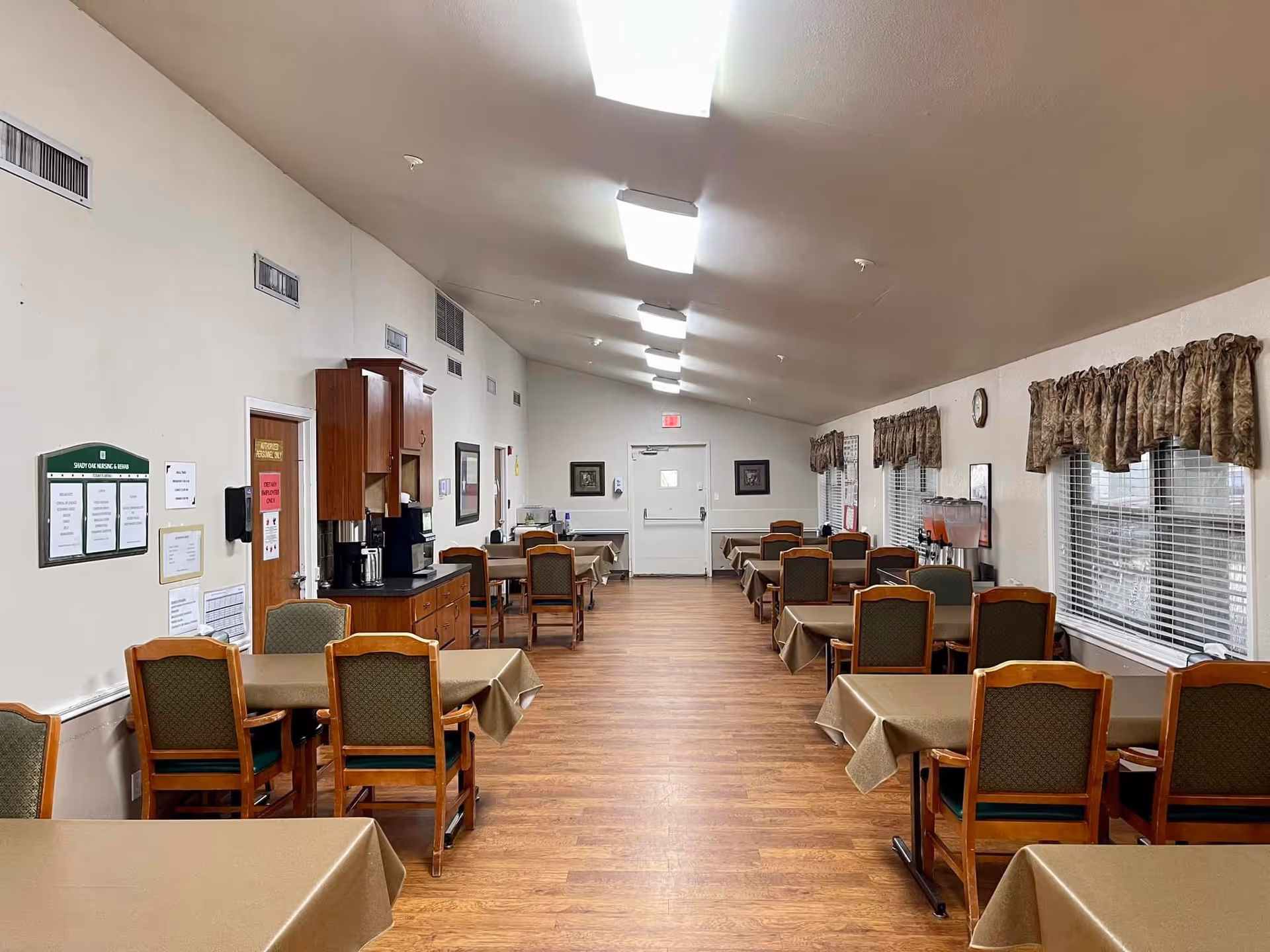 Interior view of a dining room in a senior living facility with multiple tables covered in beige tablecloths and wooden chairs with green cushions. The room has wood flooring, white walls, and several windows with patterned valances. There is a coffee station and beverage dispensers along the left wall, and fluorescent ceiling lights illuminate the space.