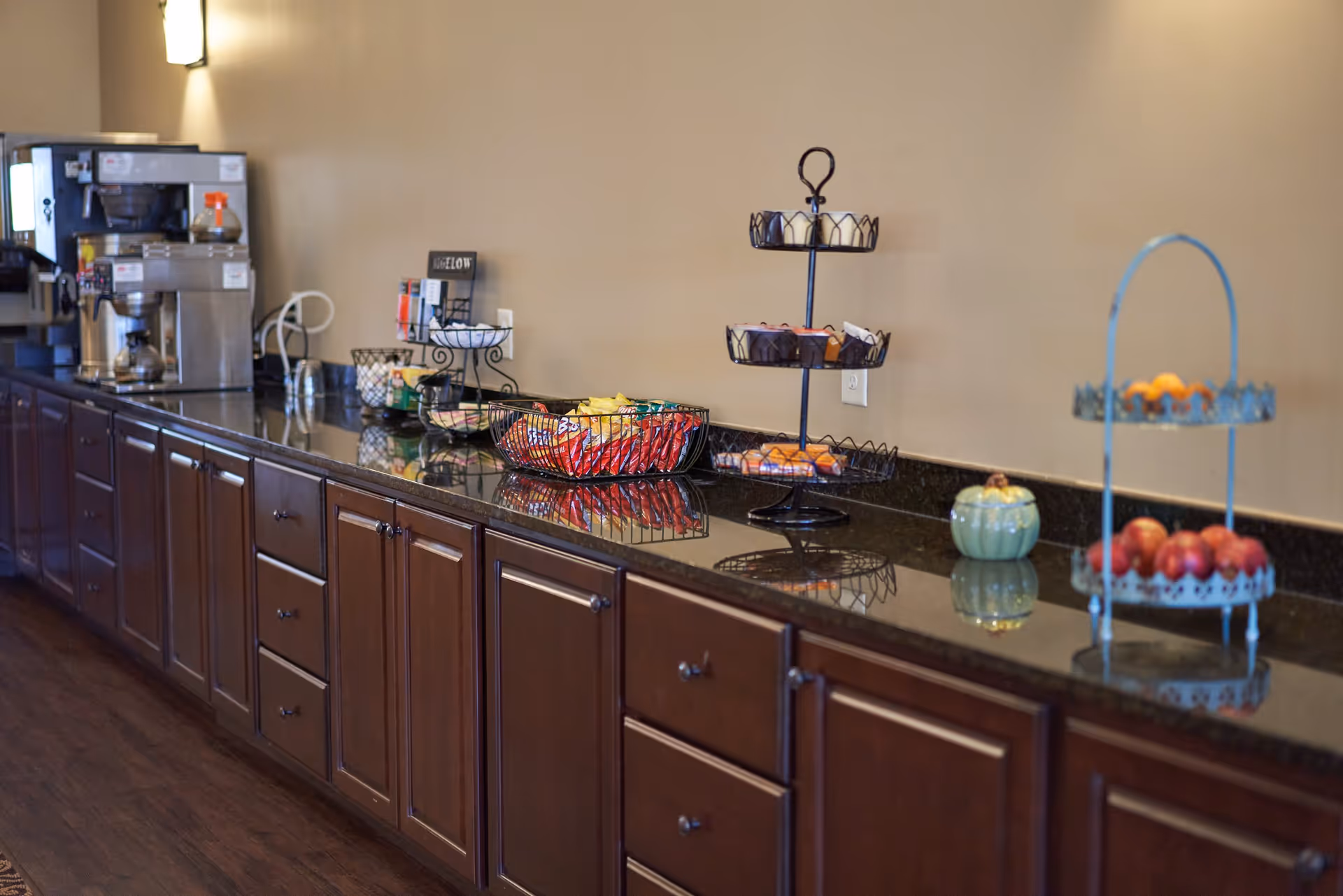 A long countertop with dark wooden cabinets underneath, featuring a coffee machine, baskets with snacks, and tiered trays holding fruit and other items against a beige wall with a wall-mounted light.
