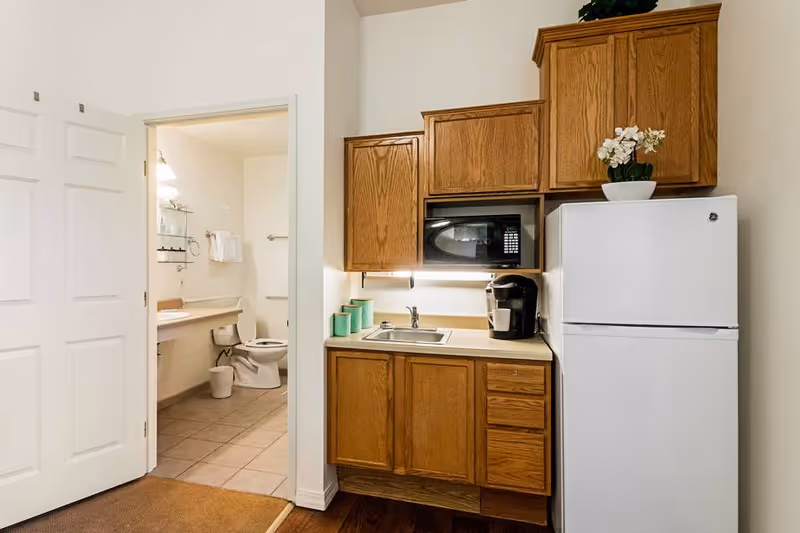 A small kitchen area with wooden cabinets, a white refrigerator, a microwave, a coffee maker, and a sink. To the left, an open door reveals a bathroom with a toilet, a sink, and a mirror with shelves above it.