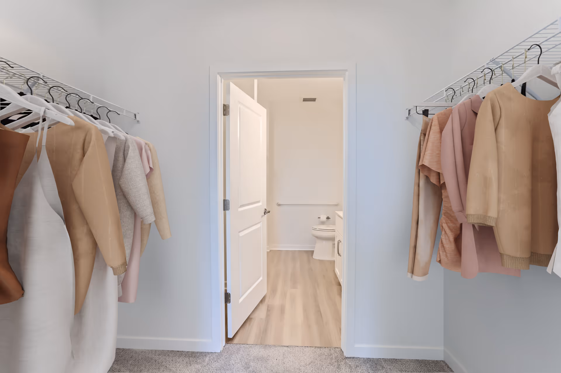 View from a walk-in closet with clothes hanging on both sides on white wire racks, looking through an open door into a bathroom with a toilet, grab bar, and vanity with cabinet. The closet has carpeted flooring and the bathroom has light wood flooring.