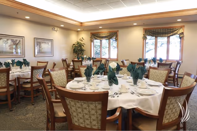 Dining room with round tables set for a meal, wooden chairs, and green folded napkins under soft ceiling lighting.