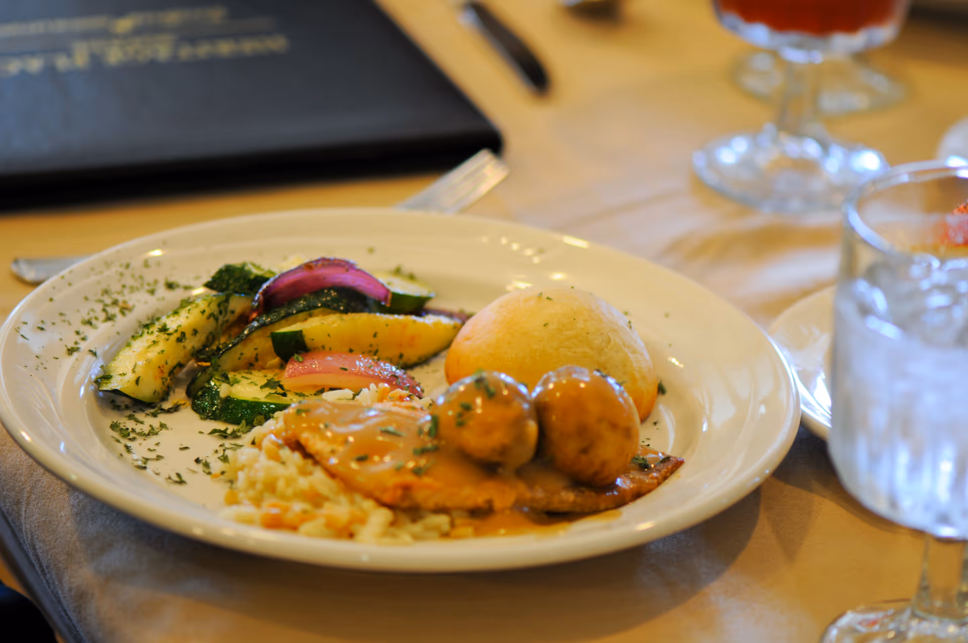 A plated meal consisting of grilled vegetables including zucchini and red onions, a bread roll, two meatballs with gravy, and a serving of rice on a white plate. The plate is on a table set with a glass of water and a glass of iced tea in the background.