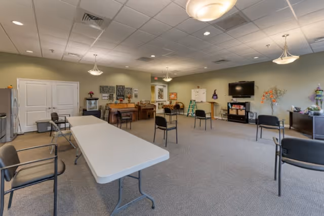 A spacious activity room in a senior living facility with chairs arranged in a circle, a long folding table, a piano, a television mounted on the wall, and various decorations including wall art and plants.