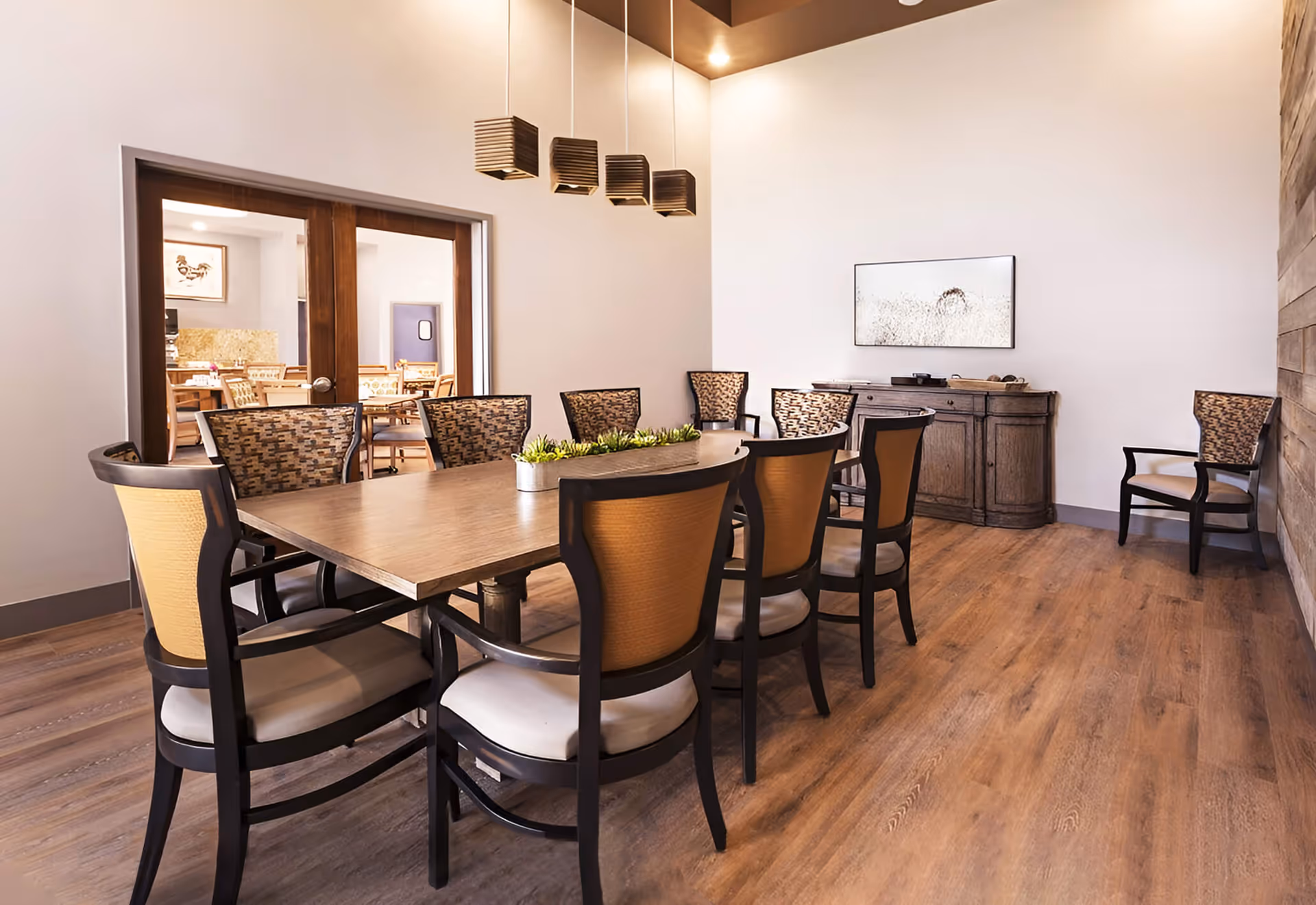 Well-lit dining room with a long wooden table surrounded by chairs, pendant lights overhead, and a sideboard against the far wall.