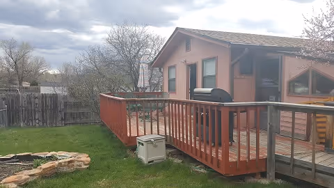 Outdoor view of a residential assisted living facility showing a wooden deck attached to a single-story building with a grill on the deck. The yard has green grass, a small stone-bordered garden area, and a wooden fence in the background under a cloudy sky.