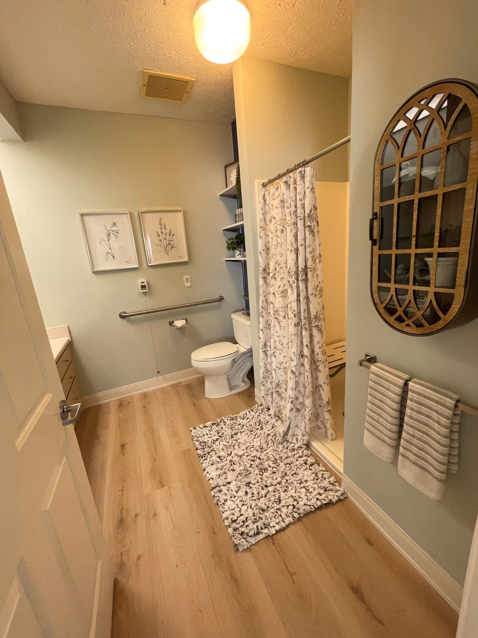 A clean and well-lit bathroom with light wood flooring, a white toilet, a shower with a floral curtain, a fluffy white and gray rug, two framed botanical prints on the wall, a grab bar next to the toilet, a towel rack with two striped towels, and a decorative wall mirror with a wooden frame.