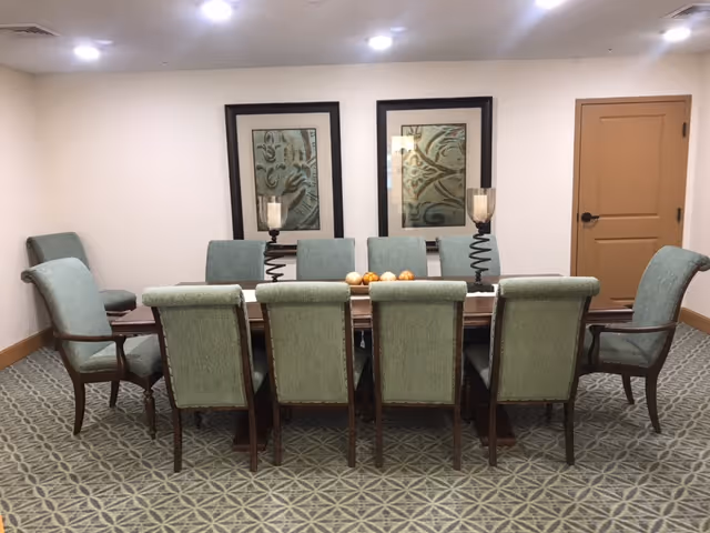 A dining room with a long wooden table surrounded by ten upholstered chairs in a light green fabric. On the table are two decorative candle holders with spiral bases and a centerpiece with decorative balls. The room has patterned carpet flooring, two framed abstract artworks on the wall, and a closed wooden door to the right.