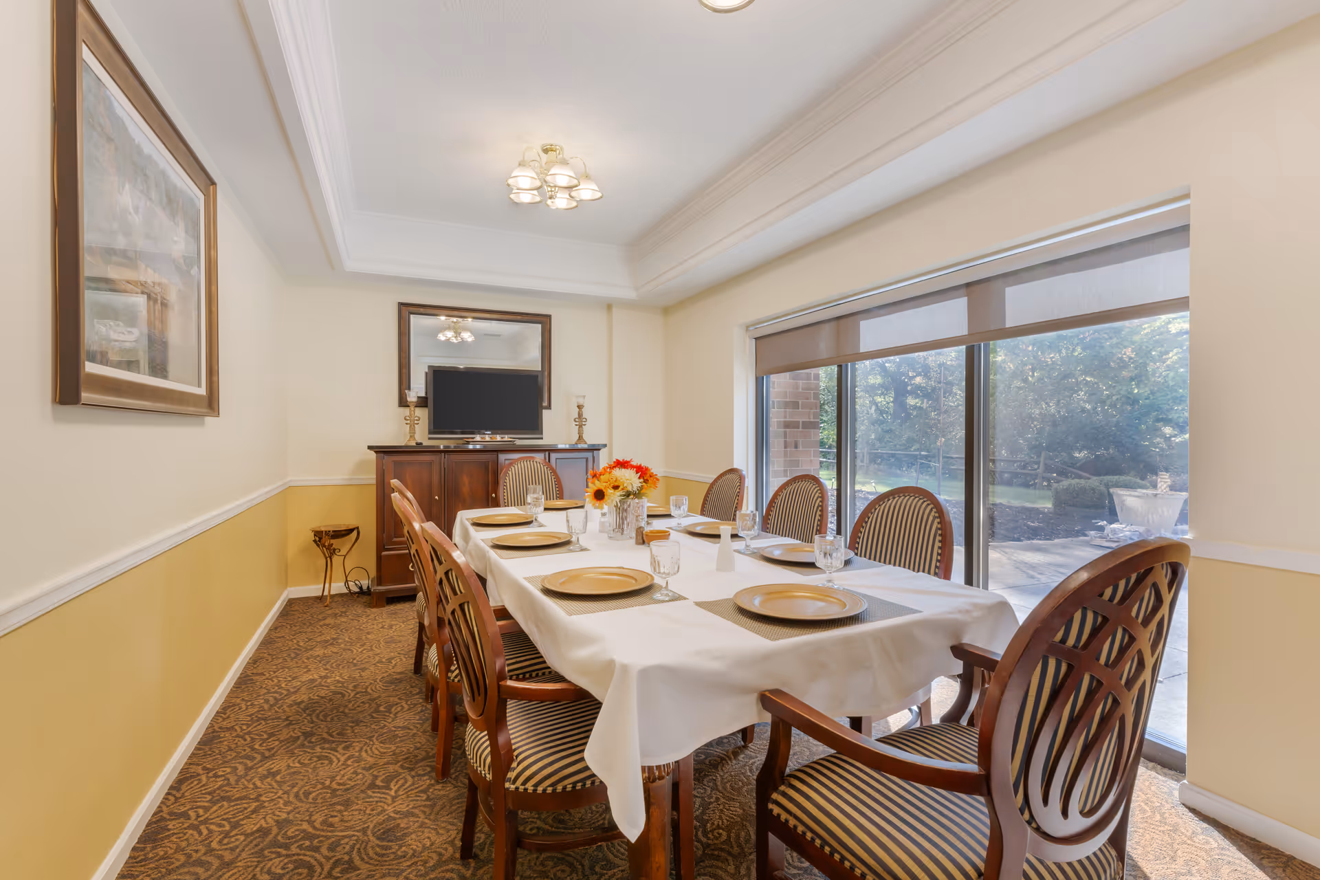 A dining room with a long table covered with a white tablecloth, set with gold plates and glassware. Eight wooden chairs with striped cushions surround the table. A sideboard with a TV and two candlesticks is against the far wall, with a large mirror above it. Large windows on the right side show an outdoor garden area.