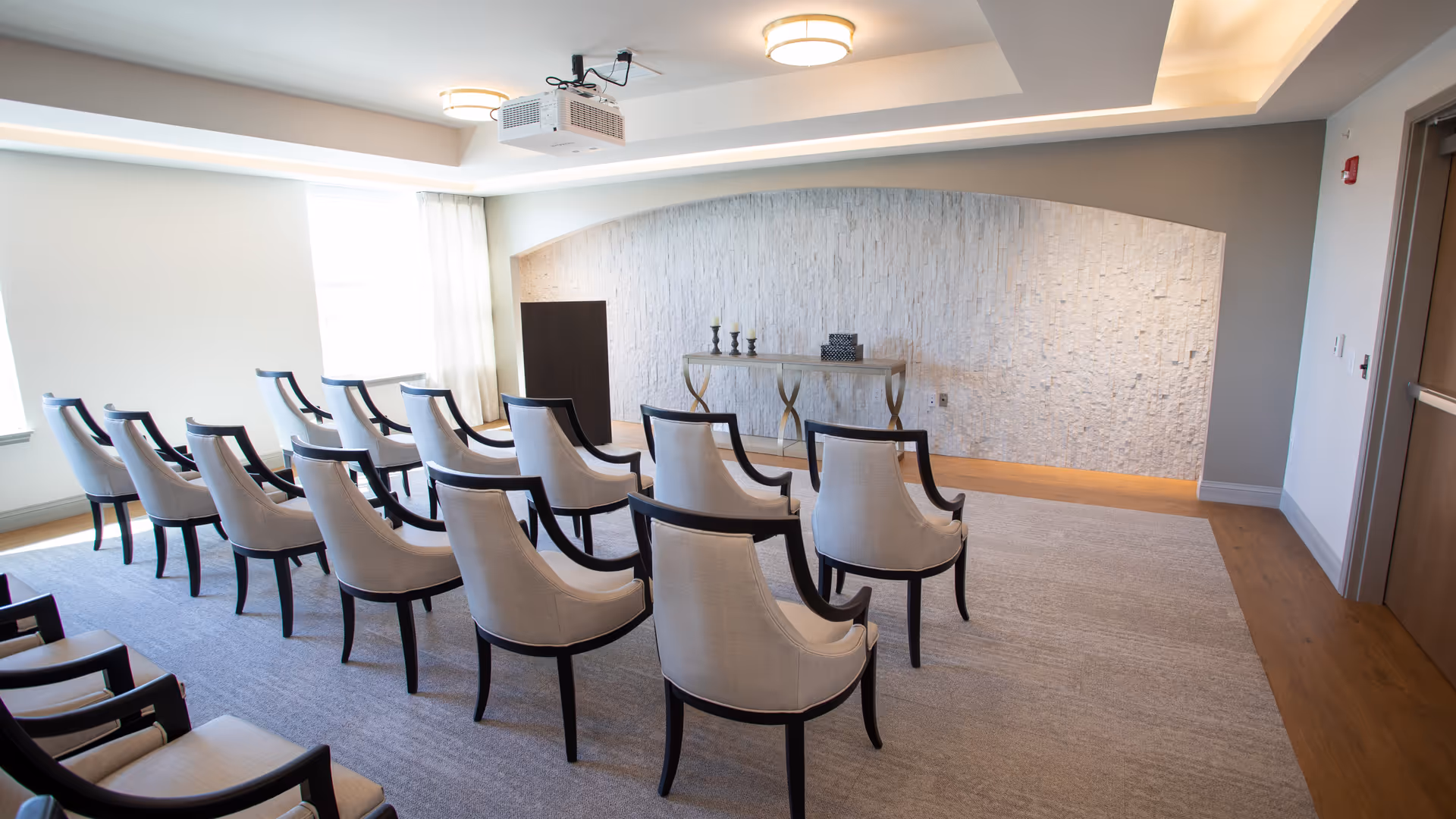 A small conference or meeting room with rows of beige upholstered chairs with dark wooden frames arranged facing a podium and a decorative table against a textured white stone wall. The room has a light carpet, wood flooring near the walls, ceiling lights, and a ceiling-mounted projector.