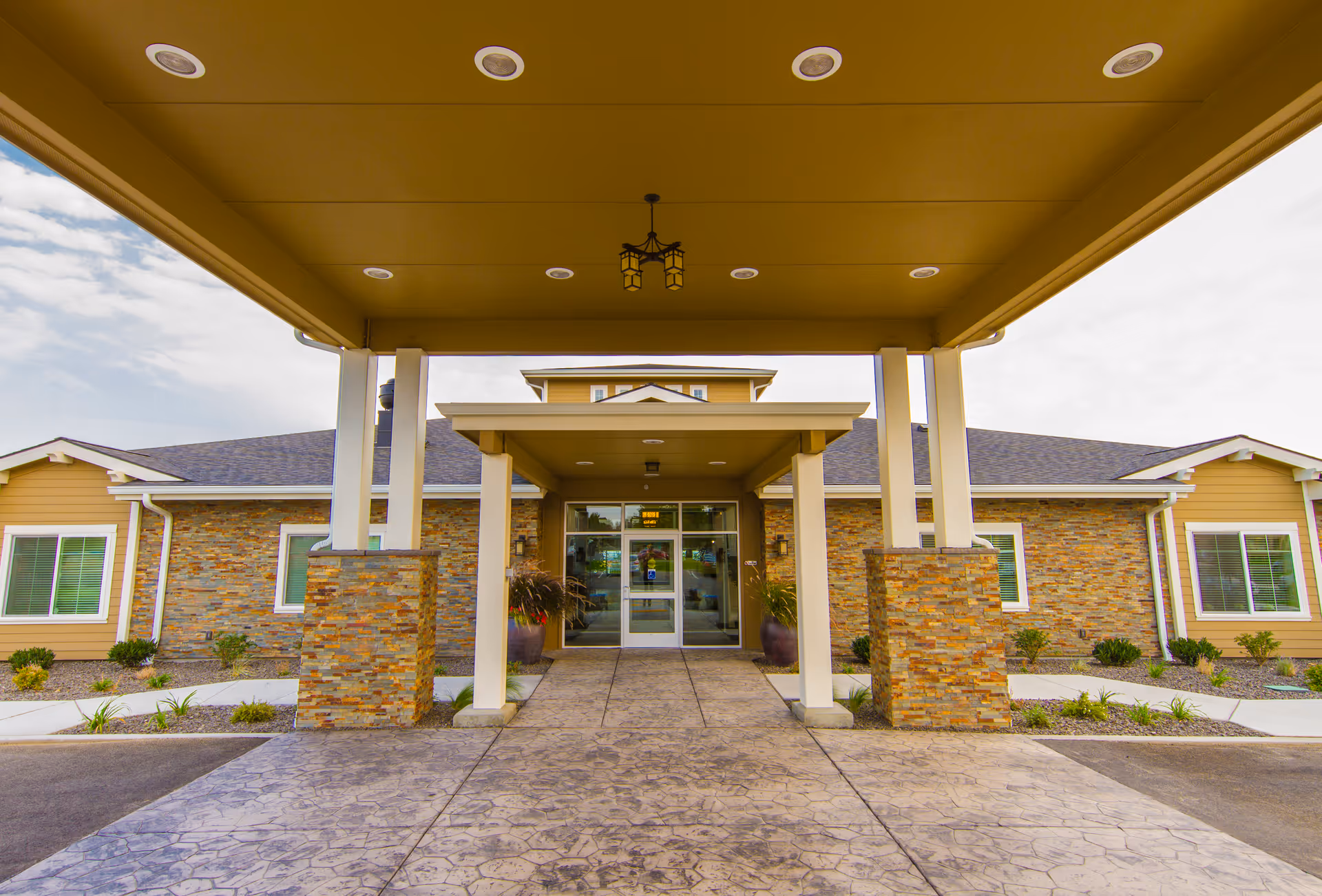 Entrance of a senior living facility with a covered driveway supported by pillars with stone bases, leading to glass double doors. The building has a stone and beige siding exterior with windows and landscaping along the walkway.