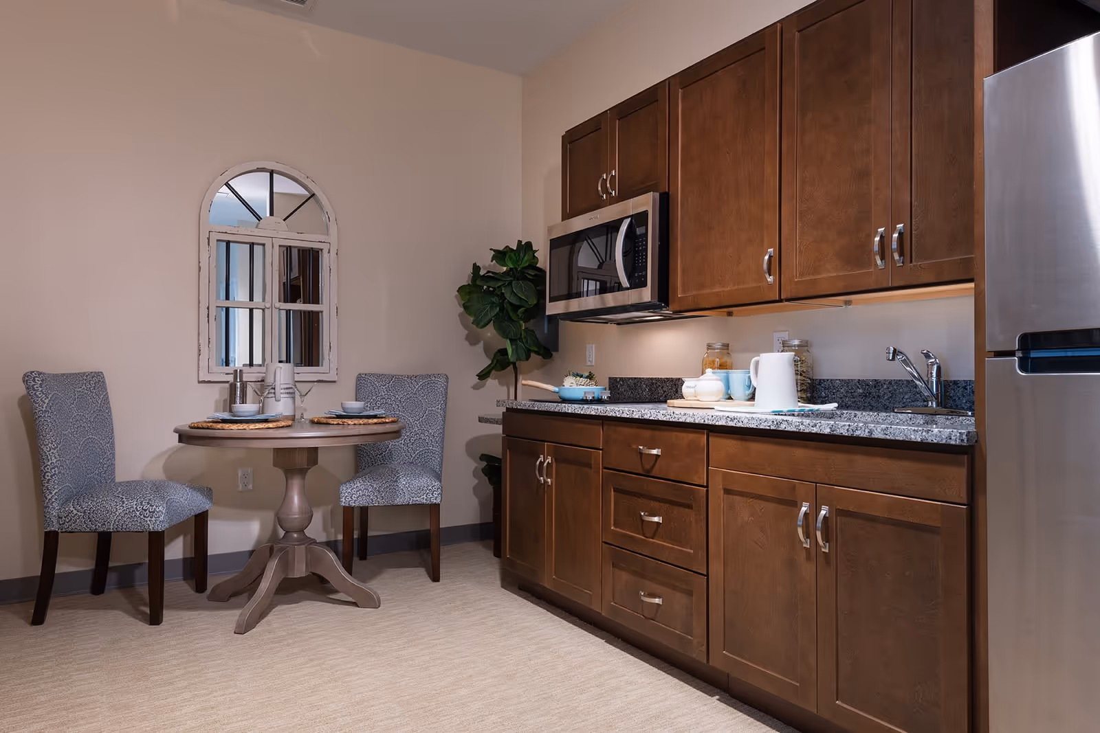Small kitchen and dining nook with wooden cabinets, granite countertop, microwave, stainless refrigerator and a round table with two upholstered chairs.