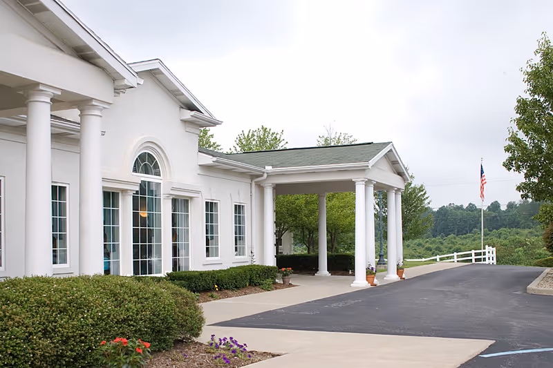 Front entrance of a white senior living facility with columns, large windows, a covered driveway, landscaping, and an American flag.