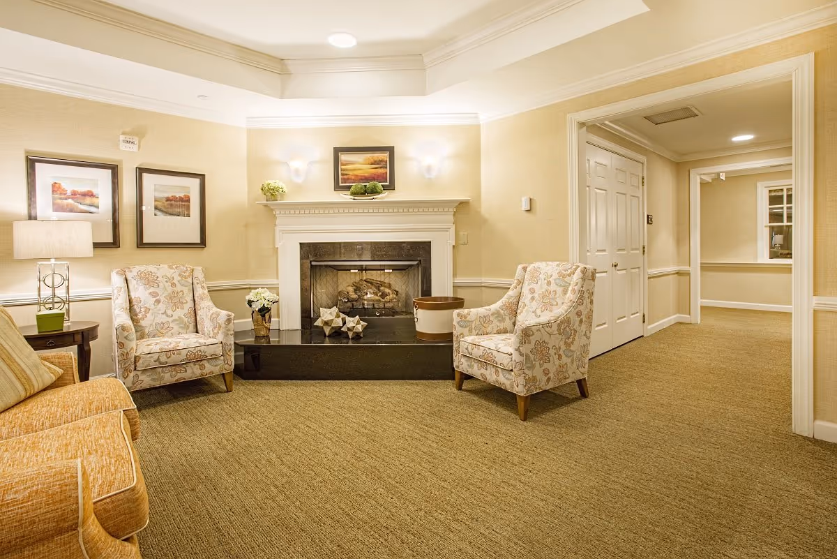 A cozy living room area with beige walls and carpet, featuring a white fireplace with decorative items on the mantel and hearth. There are two floral patterned armchairs and a yellow sofa arranged around a black coffee table. The room is softly lit by wall sconces and a table lamp, with framed landscape paintings on the wall.