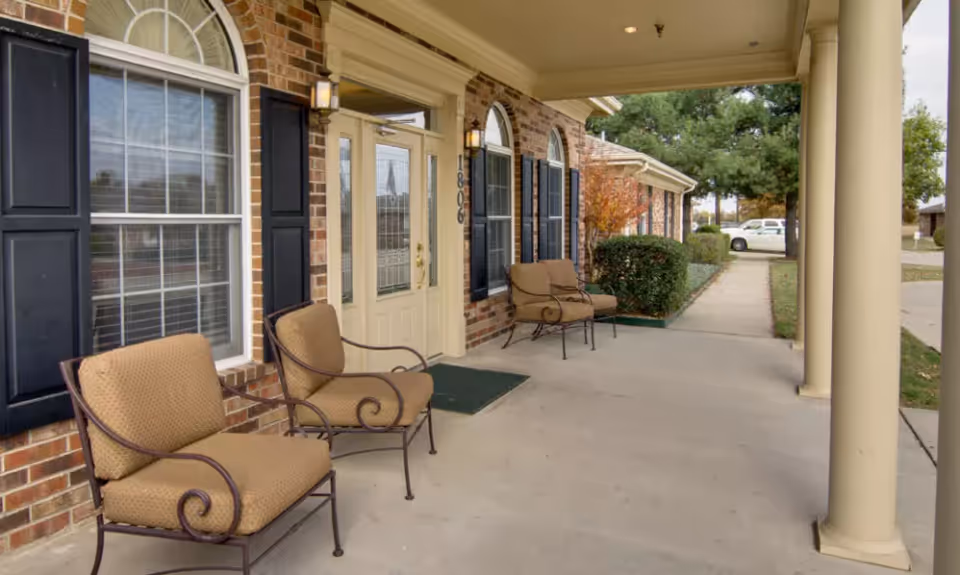 Covered front porch of a brick senior living building with cushioned chairs, columns, and a glass-paneled entrance door.
