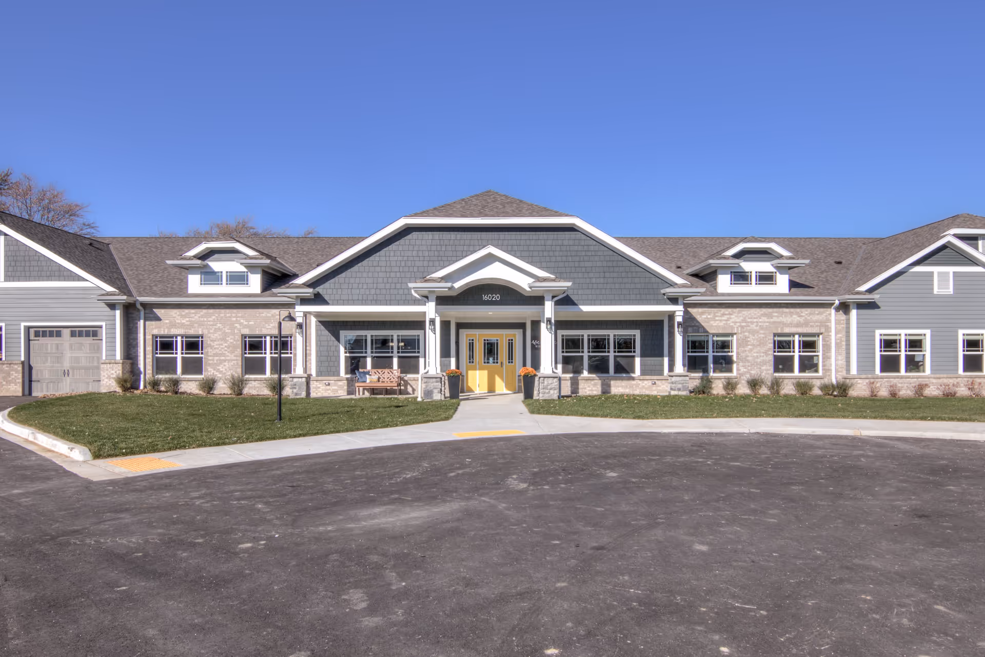 Front exterior view of a single-story assisted living and memory care facility building with gray siding, brick accents, a peaked roof, and a bright yellow double door entrance. There is a small bench and potted plants near the entrance, with a paved driveway and green lawn in front under a clear blue sky.