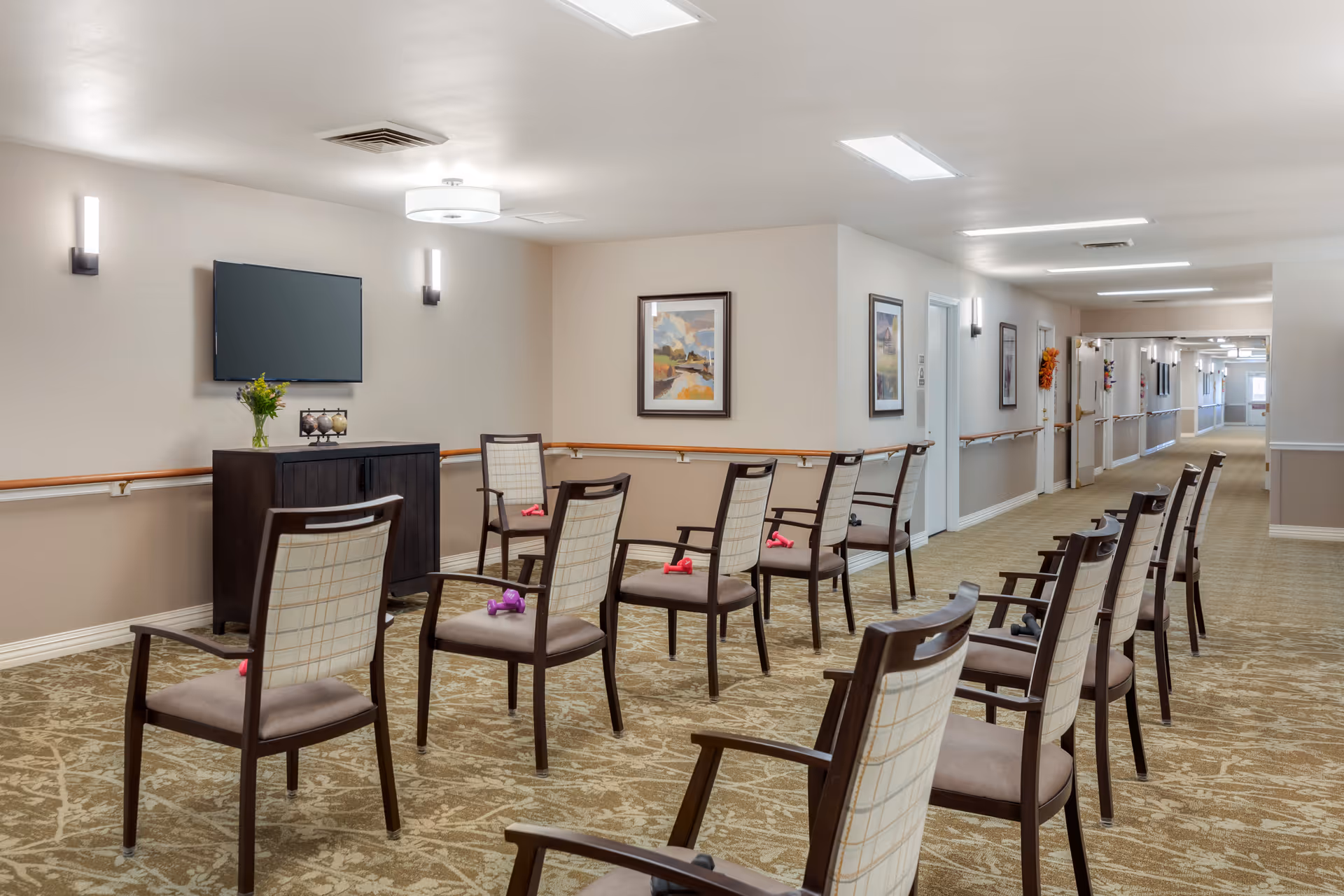 A hallway in a senior living facility with chairs arranged in two rows facing a wall-mounted TV. Each chair has a small dumbbell placed on the seat. The walls are decorated with framed paintings, and the hallway is well-lit with ceiling lights and wall sconces. There is a cabinet under the TV with a vase of flowers and decorative items on top.