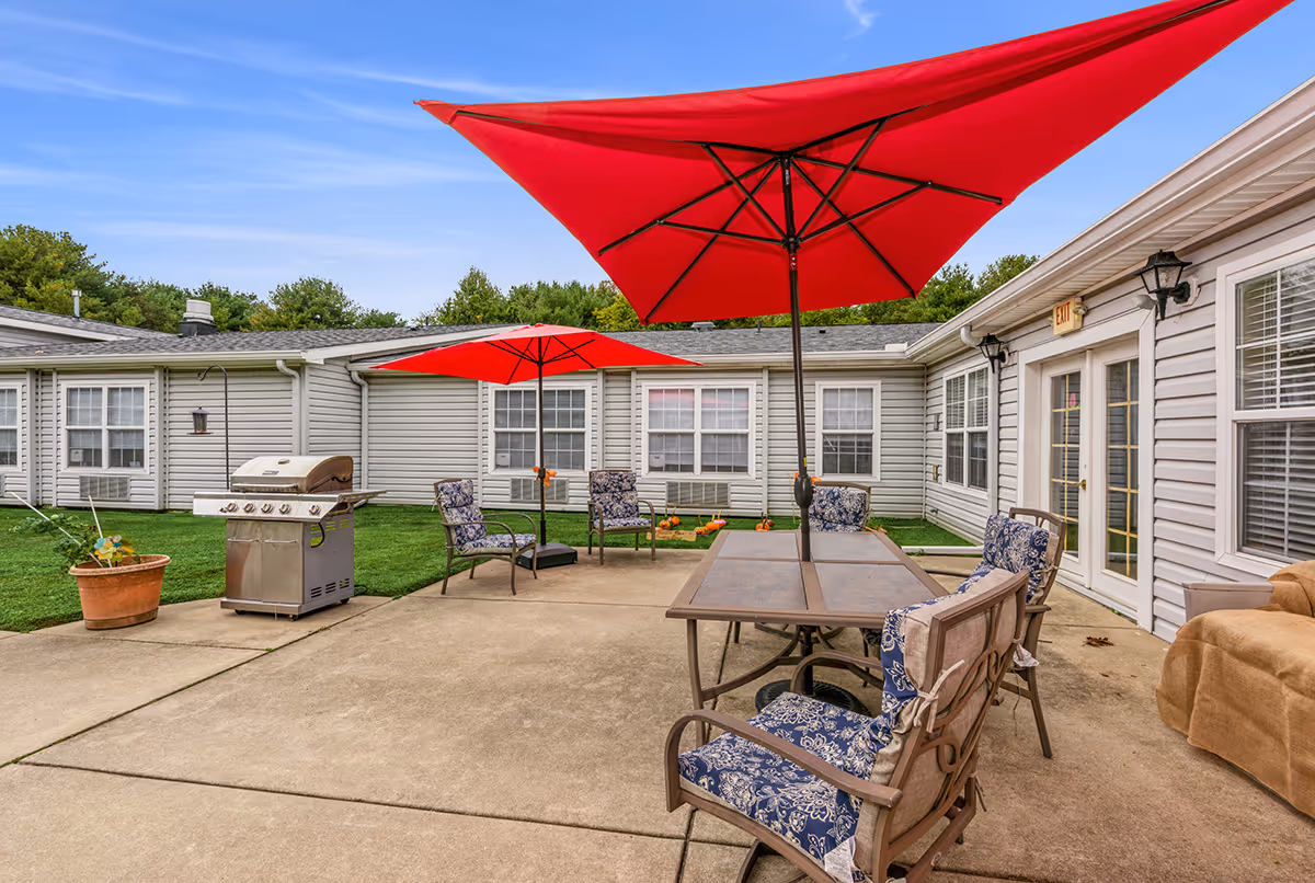 Outdoor patio area at Summit Place with concrete flooring, two red umbrellas providing shade over tables and chairs with blue patterned cushions, a stainless steel grill, potted plants, and a beige couch near the building entrance. The building has white siding and multiple windows.