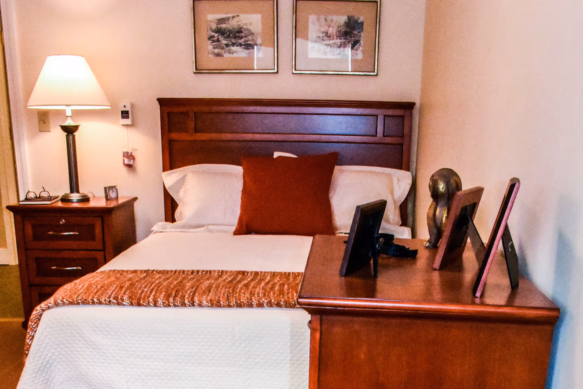 A neatly made bed with white bedding and a rust-colored throw blanket and pillow. The bed has a wooden headboard. To the left of the bed is a wooden nightstand with a lamp, a pair of glasses, and a small clock. On the right side, there is a wooden dresser with several framed photos and a decorative sculpture. Two framed pictures hang on the wall above the bed.
