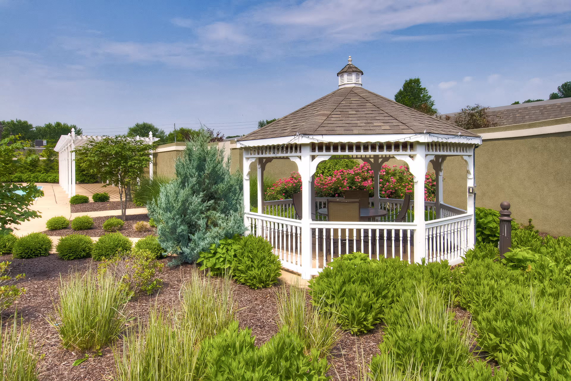 A white wooden gazebo with a shingled roof surrounded by green shrubs and plants in a garden area under a partly cloudy blue sky. There are chairs and a table inside the gazebo, and a beige wall with flowering bushes behind it.