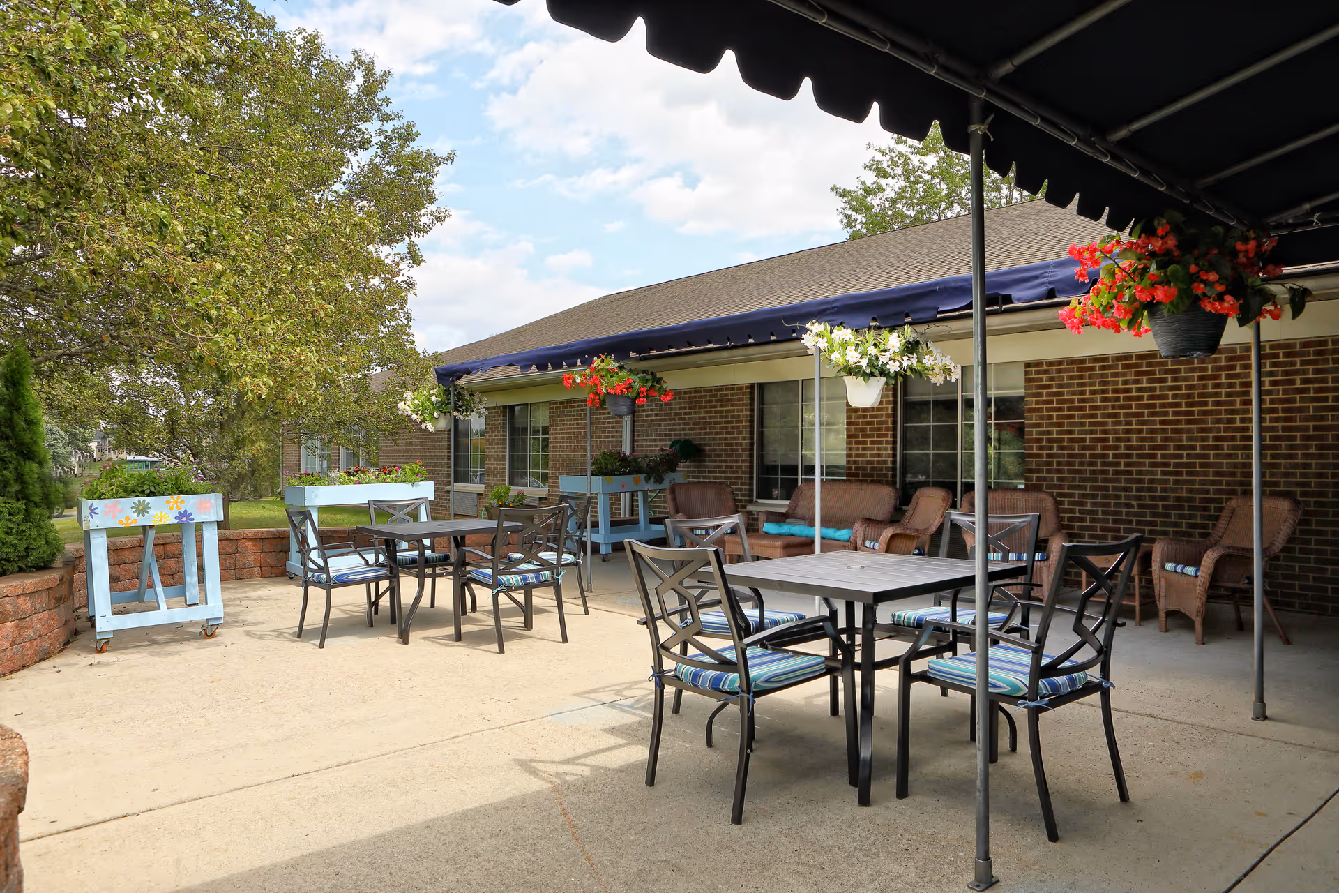 Outdoor covered patio with tables and chairs, hanging flower baskets, and a brick building in the background.