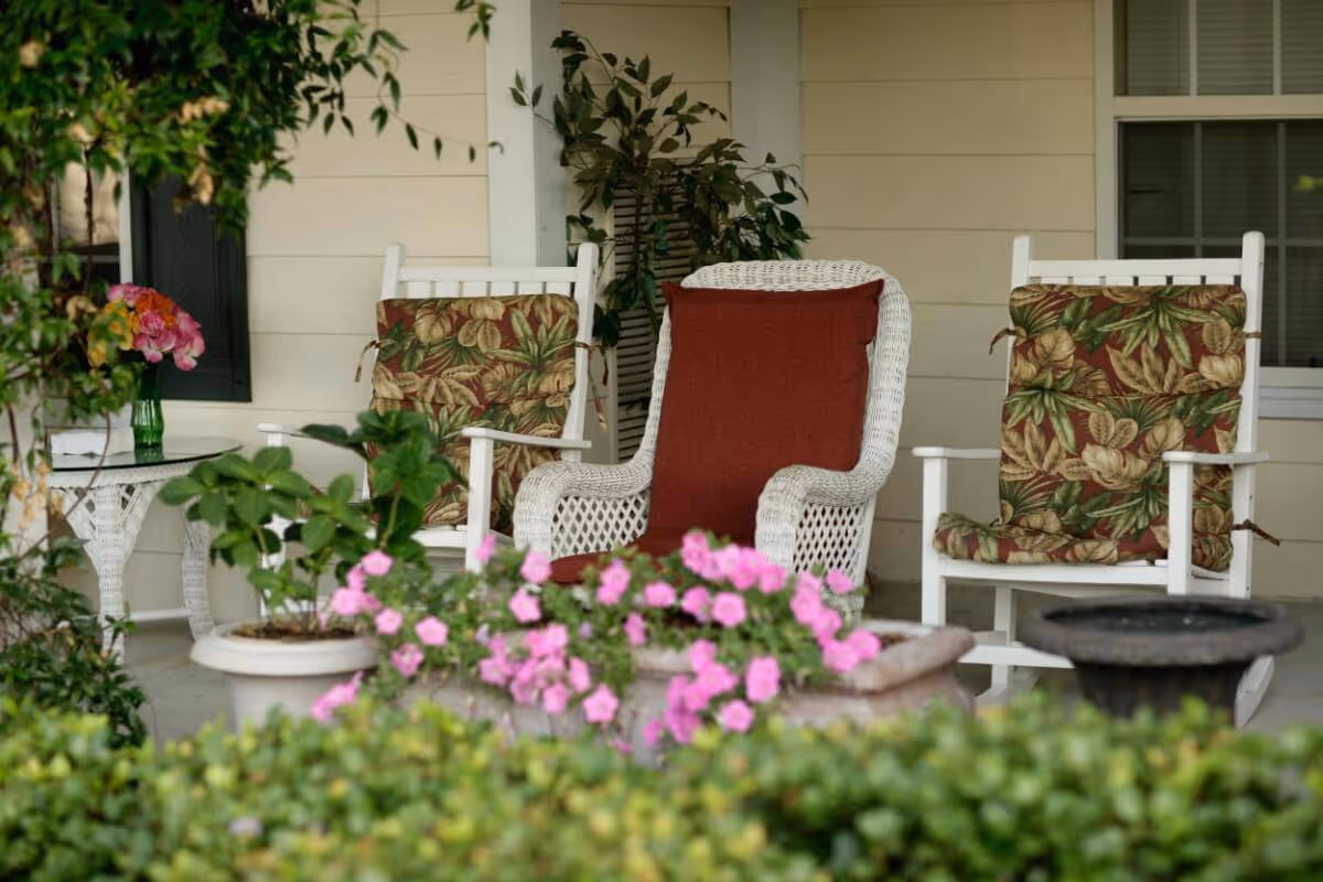 A cozy outdoor seating area with two white wooden rocking chairs featuring floral cushions and a white wicker chair with a red cushion. There are potted plants and pink flowers in the foreground, with a beige wall and window in the background.