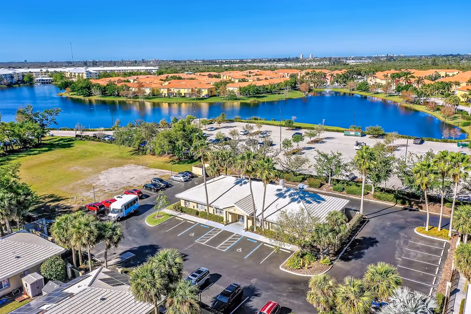 Aerial view of a senior living facility parking lot with several cars parked, surrounded by palm trees and greenery. In the background, there is a large blue lake with a fountain, and residential buildings with orange roofs. The sky is clear and blue.