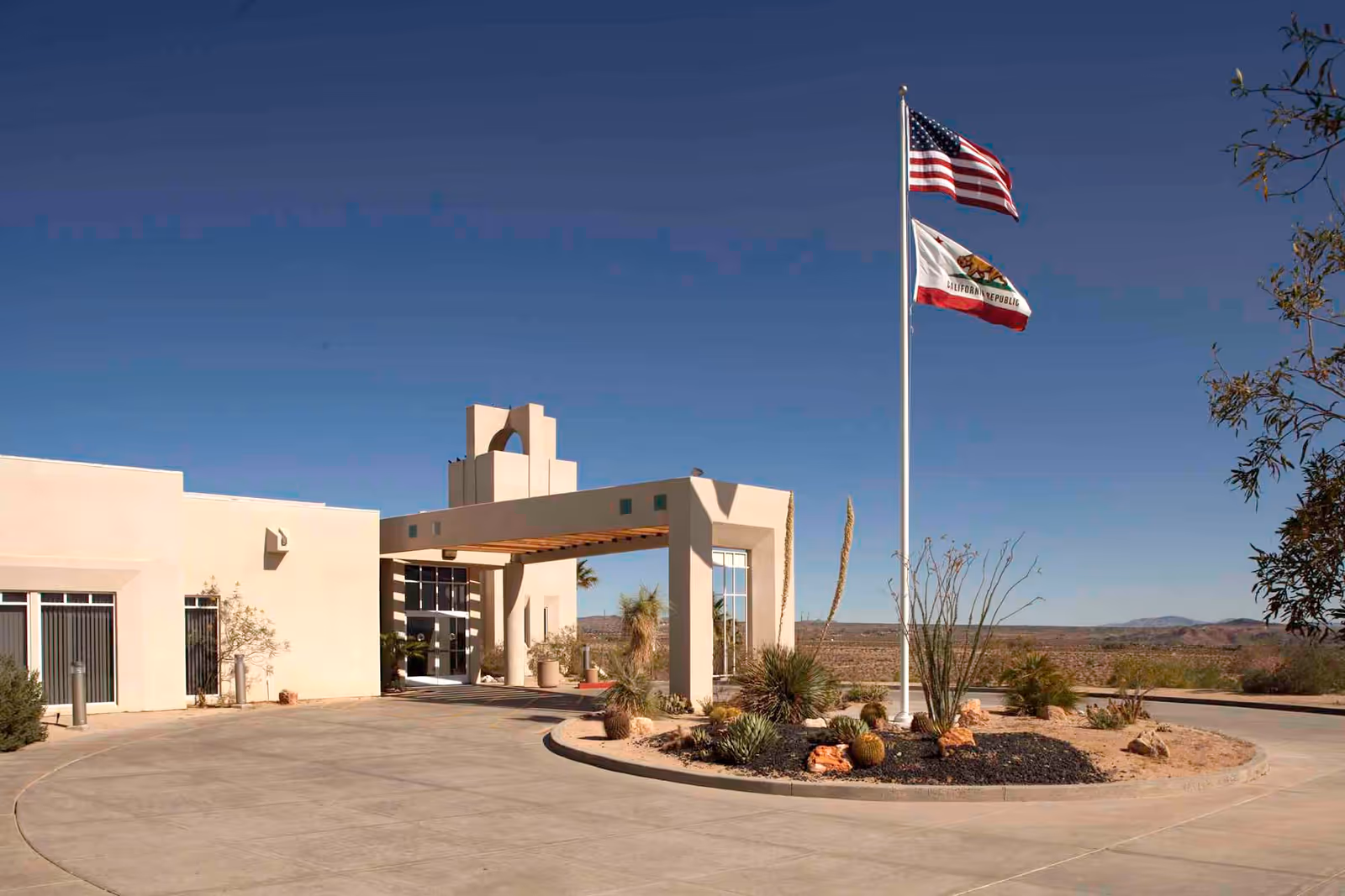 Front entrance of a cream-colored senior living building with two flagpoles, desert landscaping, and a clear blue sky.
