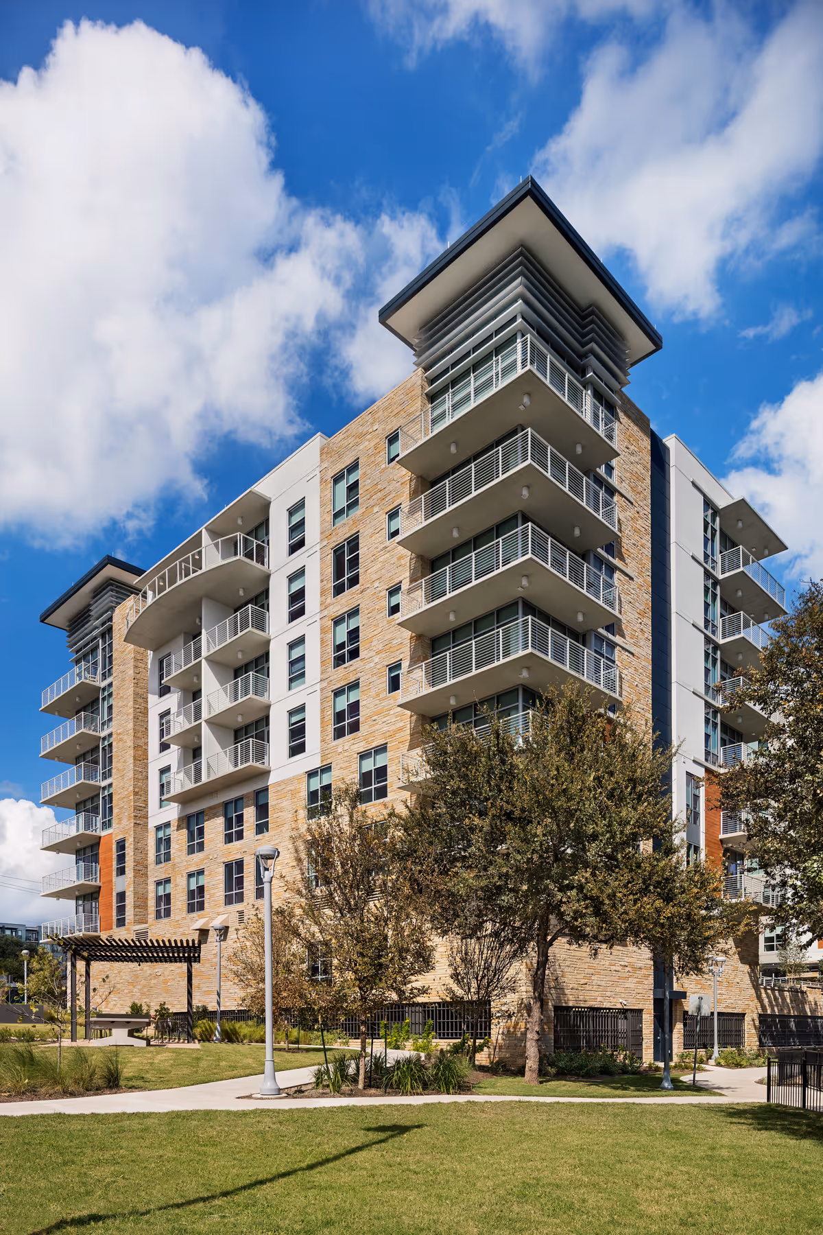 A modern multi-story residential building with balconies, surrounded by trees and a well-maintained grassy area under a partly cloudy blue sky.