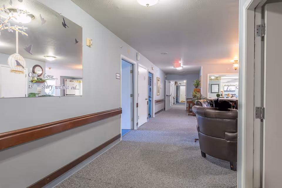 A well-lit hallway in a senior living facility with carpeted floors and light gray walls. There is a large decorative mirror on the left wall with the words 'Our Love... is story without end...' written on it. Several doors line the hallway, and at the end, there is a seating area with brown leather chairs and some plants. An exit sign is visible on the ceiling.