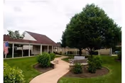 Outdoor garden area with a paved walkway leading to a seating area surrounded by bushes and a large tree, adjacent to a covered walkway and buildings in the background under a cloudy sky.